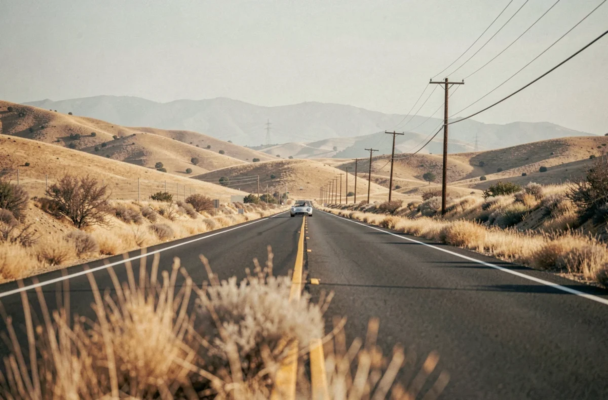 Route désertique à deux voies des collines dorées de Californie centrale avec une petite voiture de course argentée au loin, atmosphère mélancolique évoquant le dernier trajet de James Dean