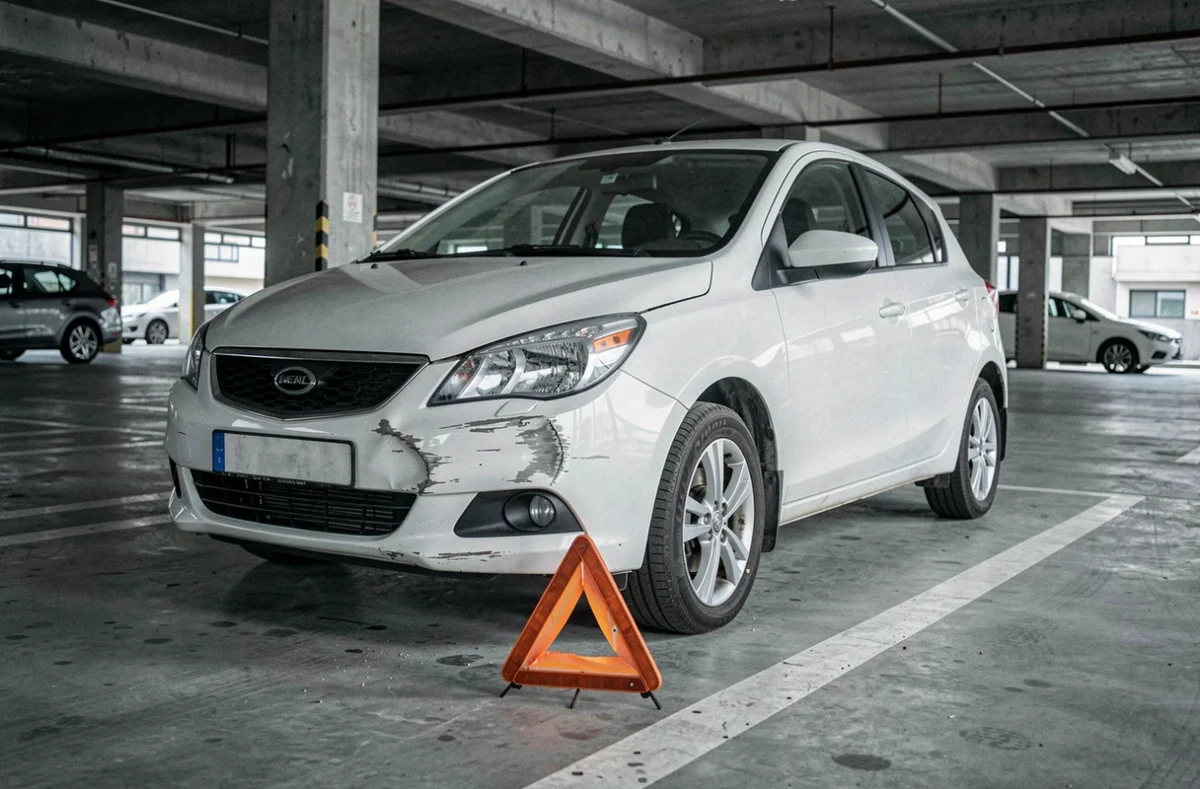 Voiture blanche accidentée avec pare-chocs endommagé dans un parking urbain, triangle de signalisation posé au sol